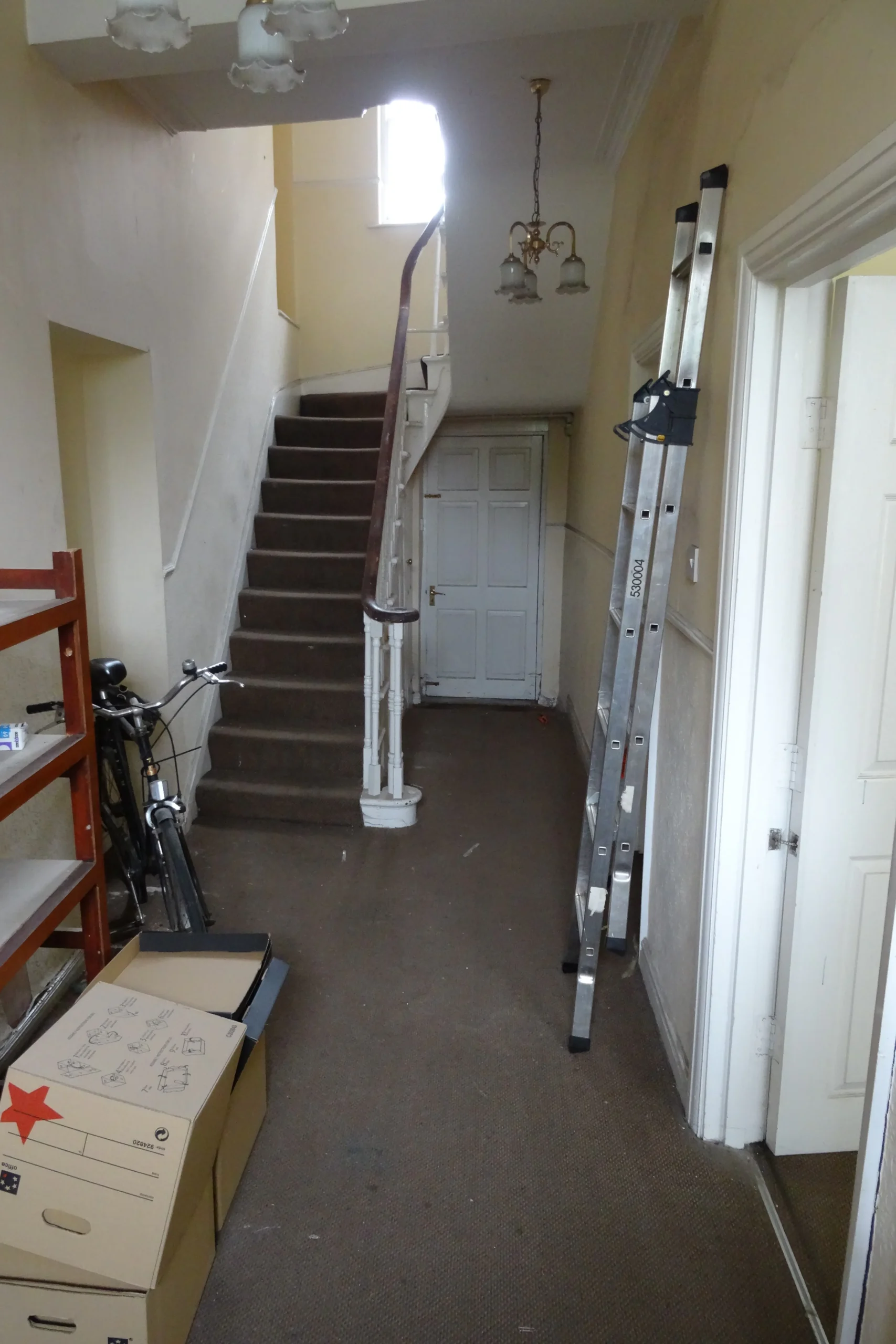 Before photograph of the original hallway of a Grade II listed townhouse in York, showing worn carpet, cluttered storage and unrestored staircase prior to full renovation