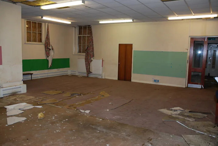 Interior of a derelict classroom at Shipton Street School with debris on the floor, boarded-up windows, and 20th-century suspended ceilings waiting for sympathetic conversion.