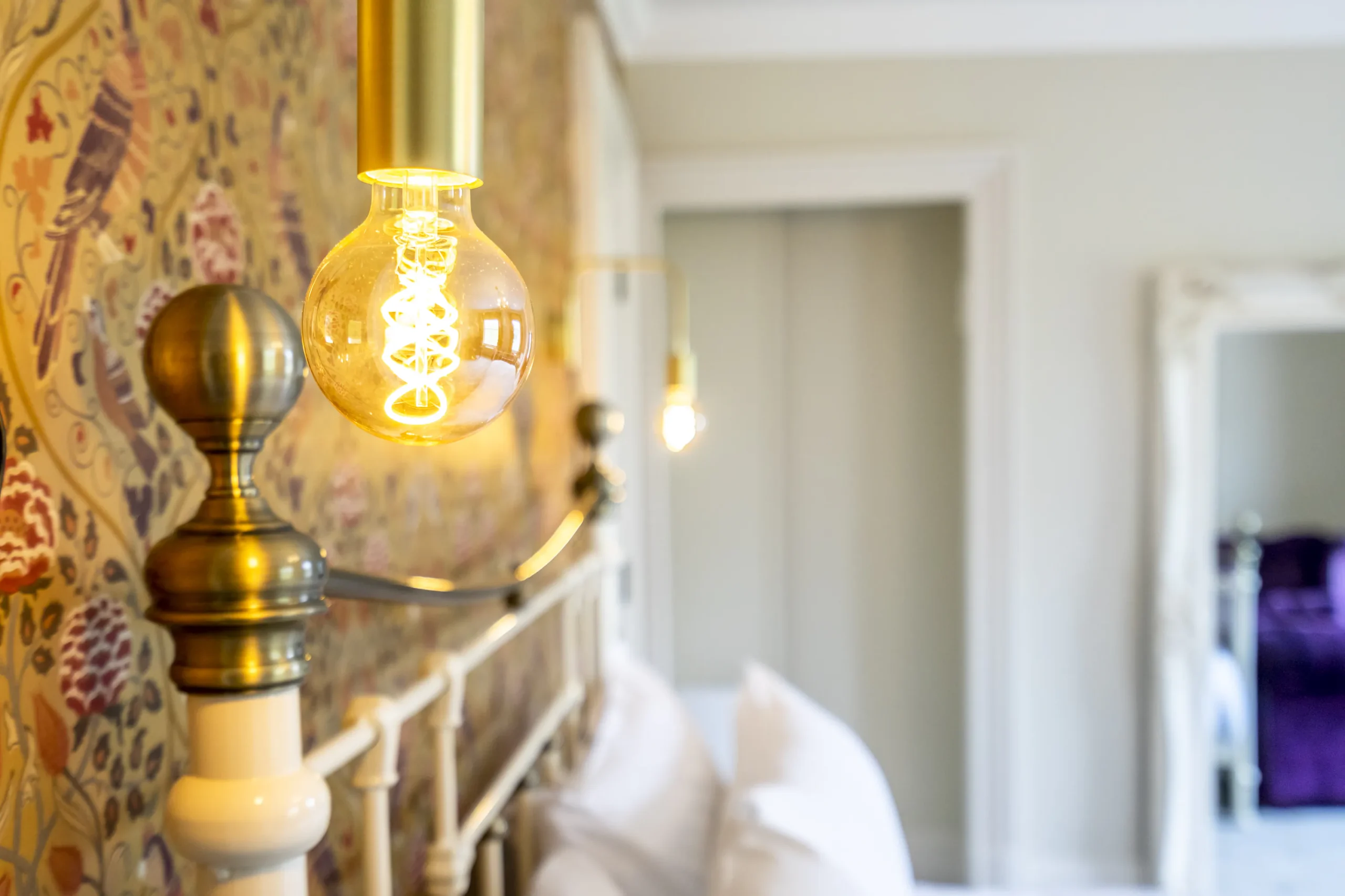Close-up bedroom detail in a Grade II listed townhouse in York featuring a brass wall-mounted Edison bulb light against ornate floral wallpaper with an iron bedstead in the foreground