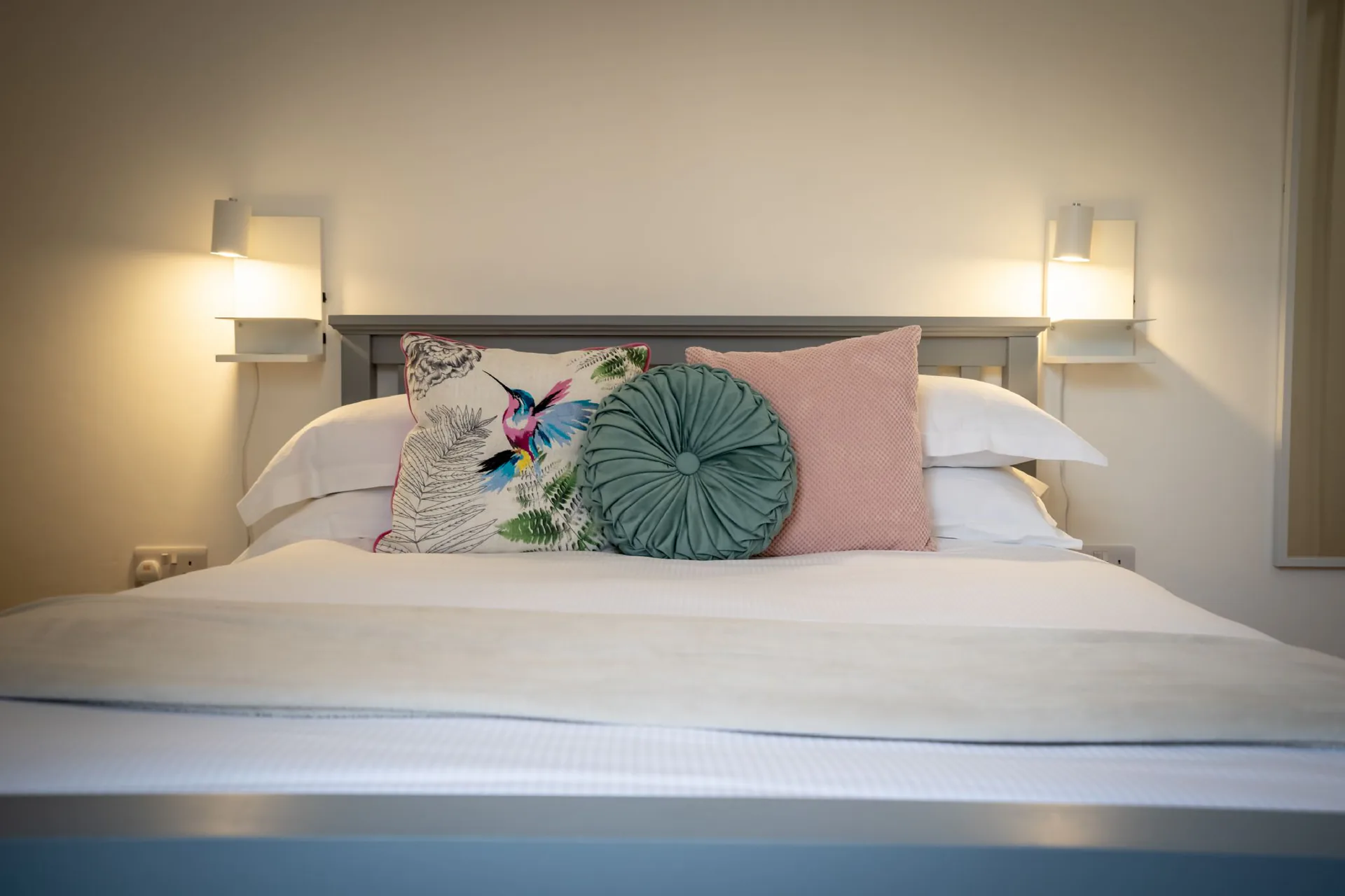 Grey-painted wooden bed headboard with white linen, hummingbird cushion and teal scatter cushion, flanked by wall-mounted reading lights in a converted Victorian listed building apartment, York
