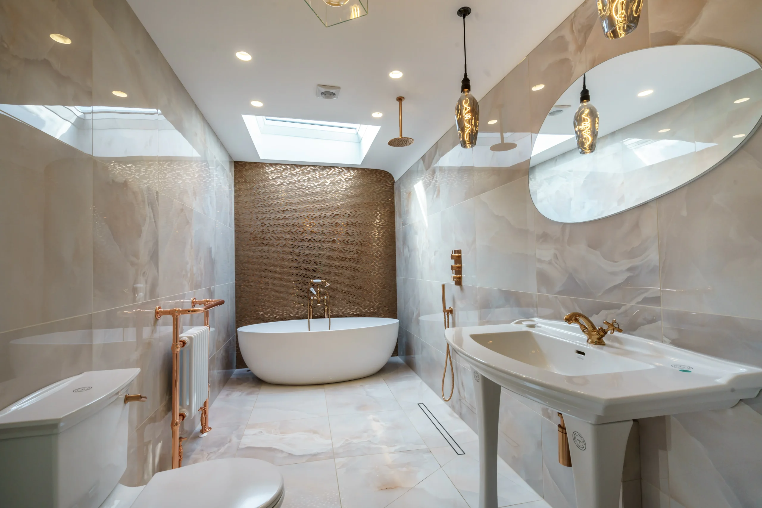 Wide-angle view of the luxury bathroom in a refurbished Grade II listed townhouse in York, featuring a freestanding oval bath, copper mosaic feature wall, rooflight, pedestal basin with brass taps, copper towel rail, Edison pendant lights and large format marble-effect floor and wall tiles