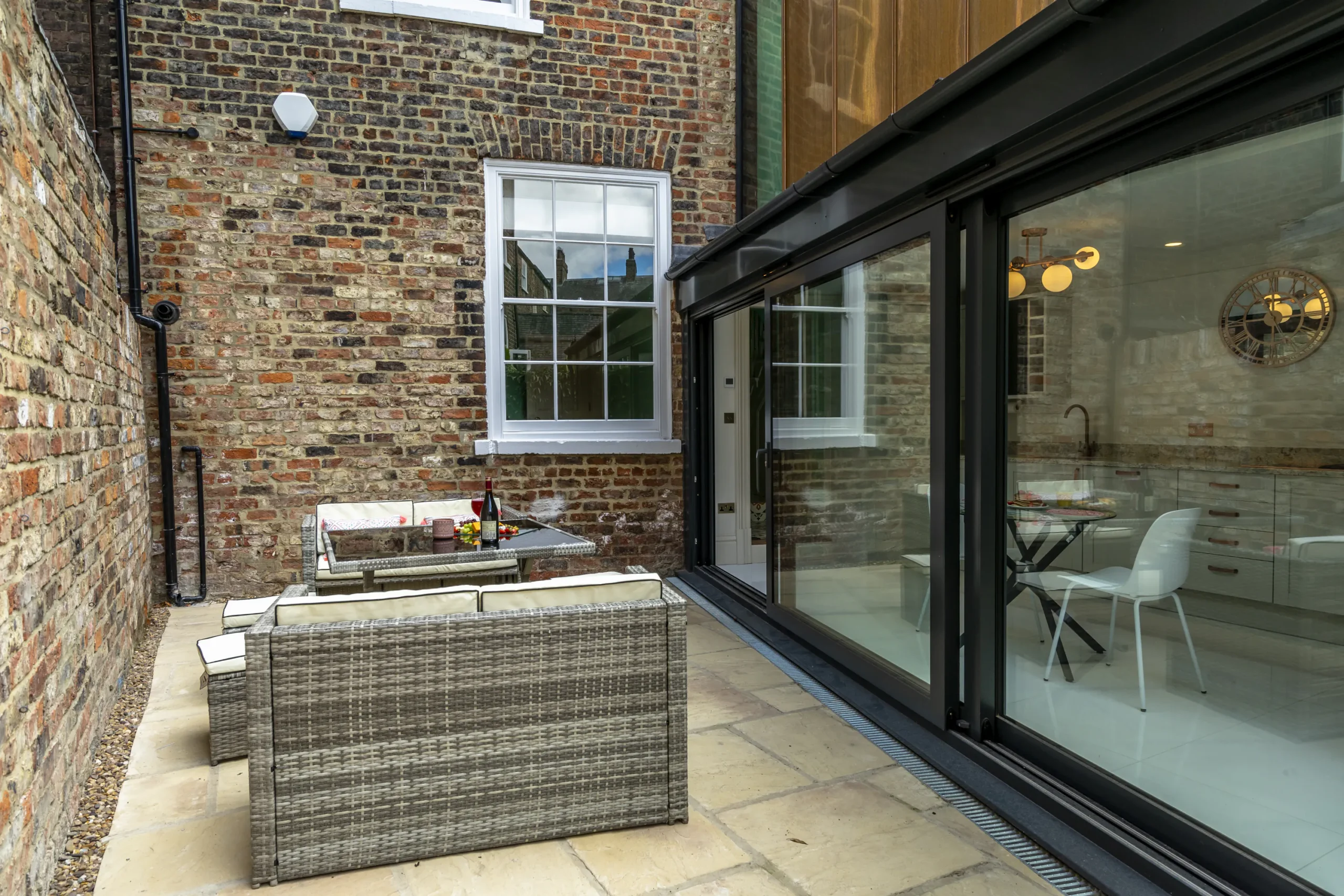 Contemporary rear extension to a Grade II listed townhouse in York featuring slim-profile aluminium sliding glass doors opening onto a sandstone patio with rattan garden furniture and exposed historic brickwork