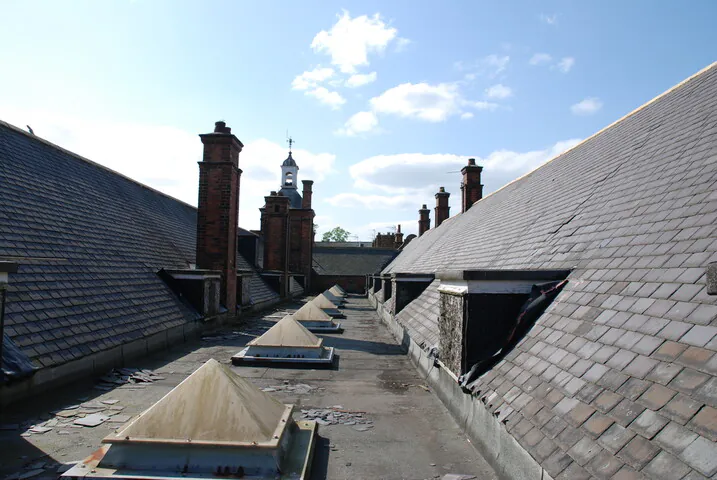 A view along a derelict flat roof section between two slate-covered pitched roofs, featuring several original Victorian red brick chimneys and pyramid skylights.