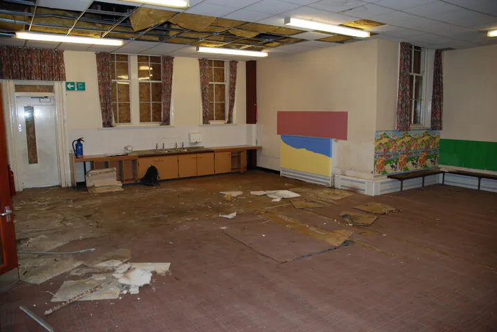 A dilapidated school room showing a dated kitchenette, peeling paint, and a partially collapsed suspended ceiling, revealing the original building's structural scale.