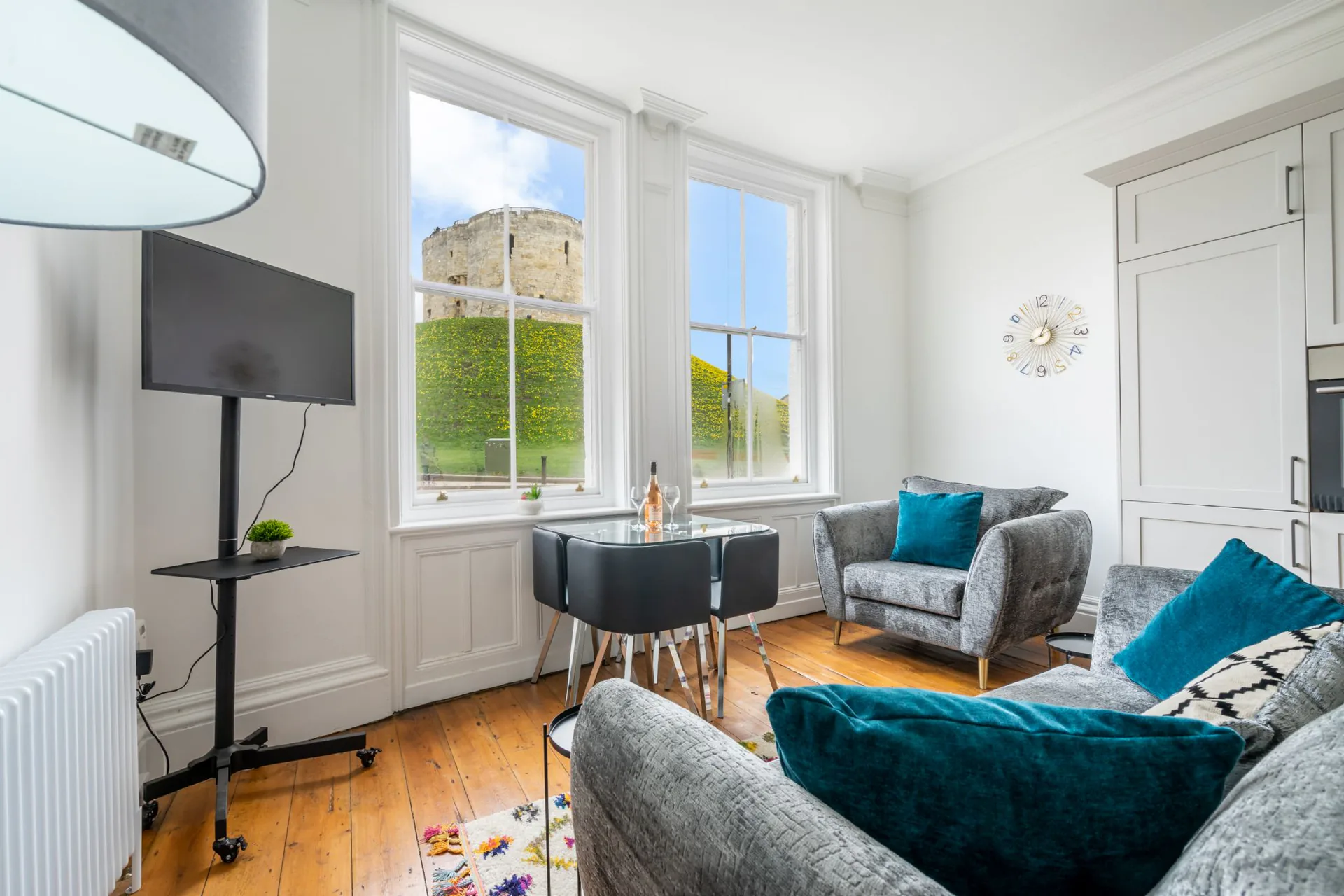 Open-plan living and dining area with twin tall sash windows framing Clifford's Tower, grey velvet sofas, glass dining table and original pine floorboards in a Grade II listed apartment conversion, Tower Street York