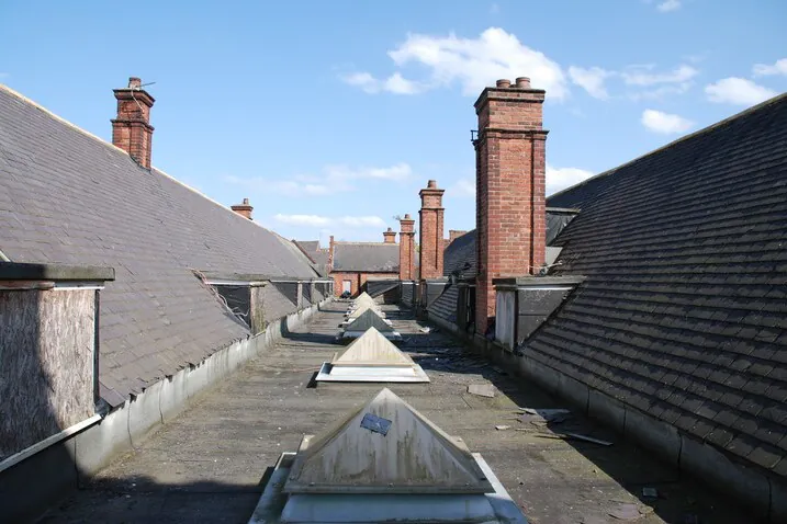 A wide perspective of the original Victorian school roof at Bootham Green, showing weathered slate tiles, brick chimneys, and boarded-up windows prior to the listed building conversion.