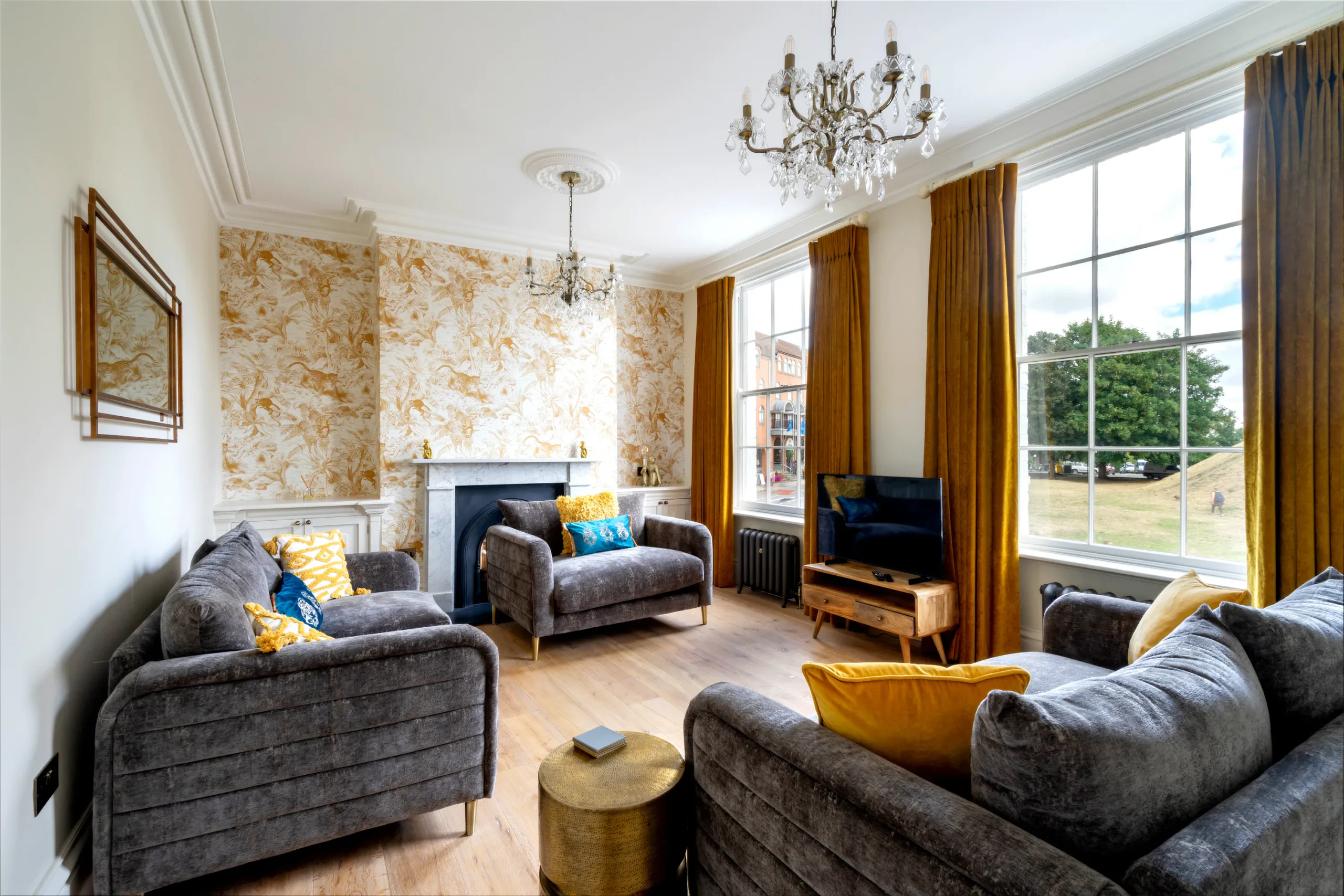 Elegant sitting room in a restored Grade II listed townhouse on Tower Street York, with original marble fireplace, ornate plaster ceiling rose, crystal chandelier, gold botanical wallpaper, velvet sofas and twin sash windows overlooking Clifford's Tower mound