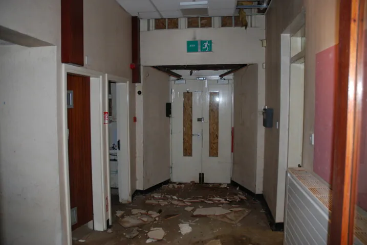 A cluttered, narrow corridor within the original 1890 school building with an exit sign and debris, showing the building's state before modern residential remodeling.