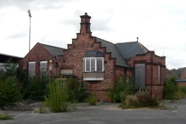 The exterior of the Victorian red brick school building featuring stepped gables and boarded windows, heavily overgrown with weeds before the start of the listed building conversion.