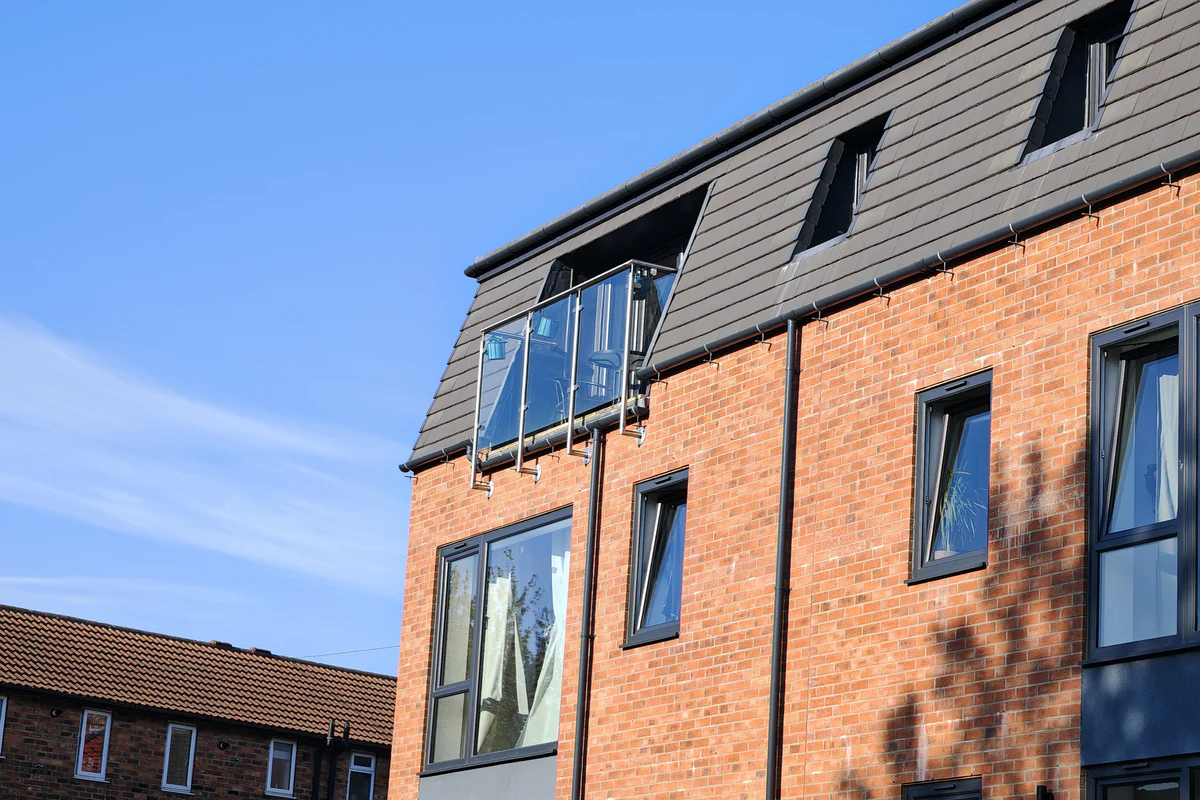 Detail of the west-facing cycleway elevation at Hawthorn Street Apartments, York, with a glass and stainless steel Juliet balcony projecting from the slate mansard roof, aluminium windows and red-brick facing contextual to the surrounding Victorian streetscape.