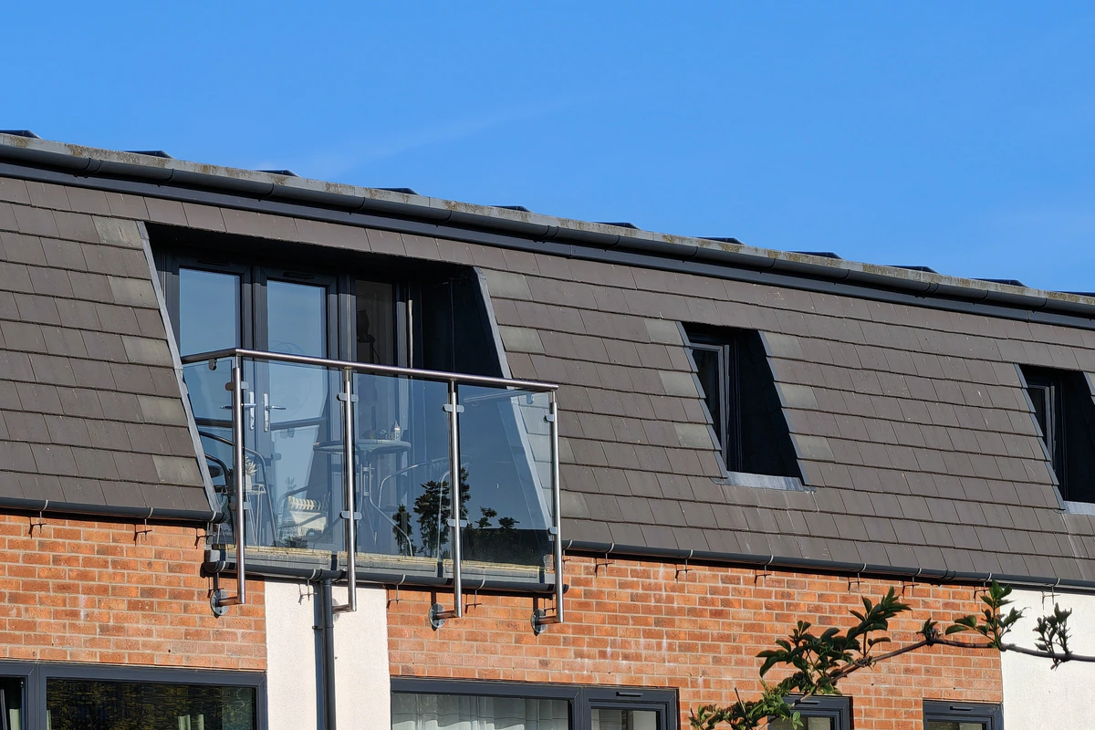 Close-up of the stainless steel and glass Juliet balcony with flanking angled dormer rooflights recessed into the dark slate mansard roof at Hawthorn Street Apartments, Layerthorpe York — contemporary detailing referencing the site's industrial workshop heritage.