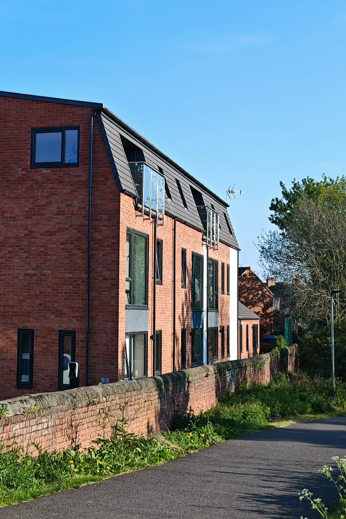 Full west elevation of Hawthorn Street Apartments running along the Foss Islands traffic-free cycleway, Layerthorpe York, showing the L-shaped contemporary new-build with multiple Juliet balconies, slate mansard roof and red-brick facade in the early evening light.