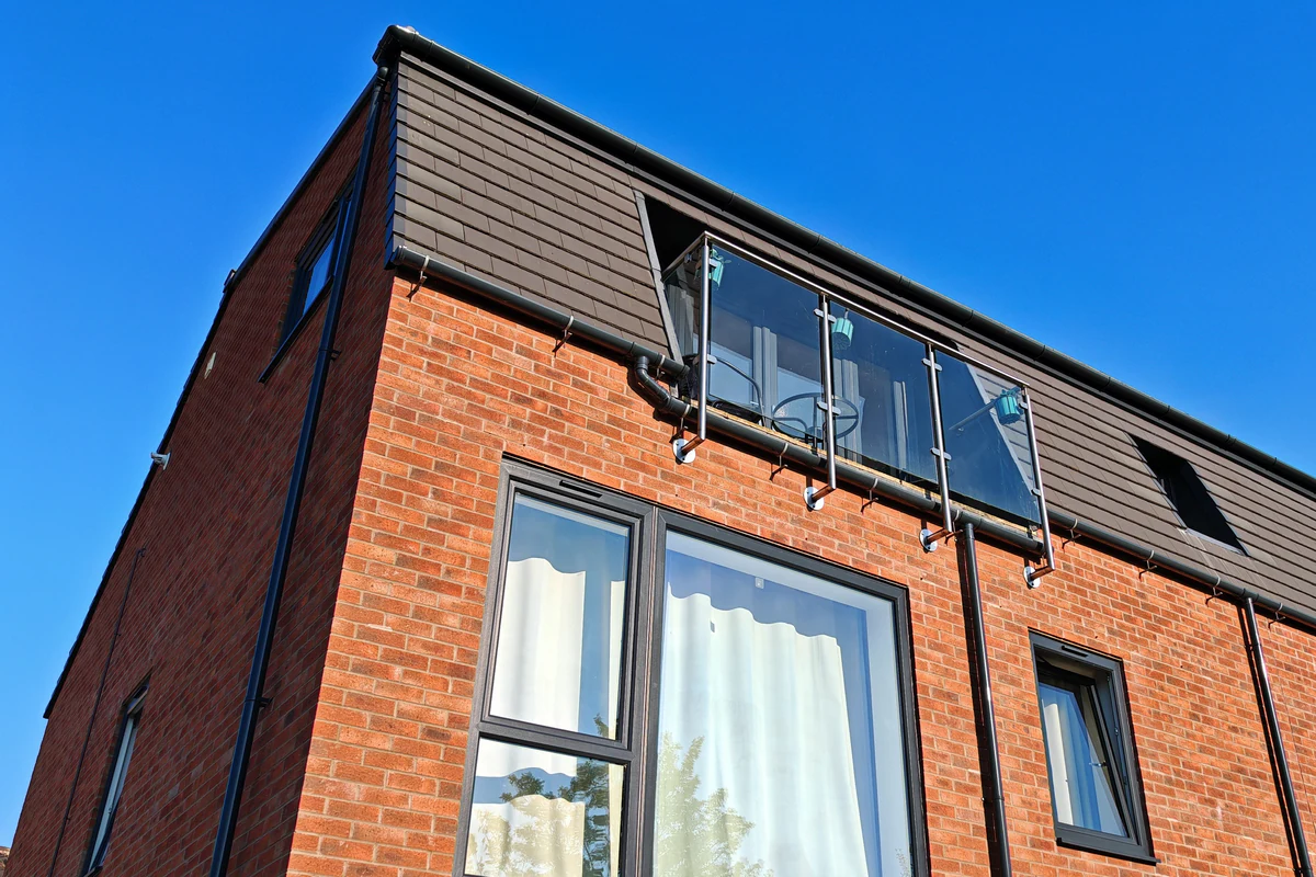 Low-angle corner view of Hawthorn Street Apartments, York, highlighting a projecting glass Juliet balcony at mansard roof level, large aluminium-framed windows and red-brick facing — contemporary residential architecture by Fining Associates chartered architects.