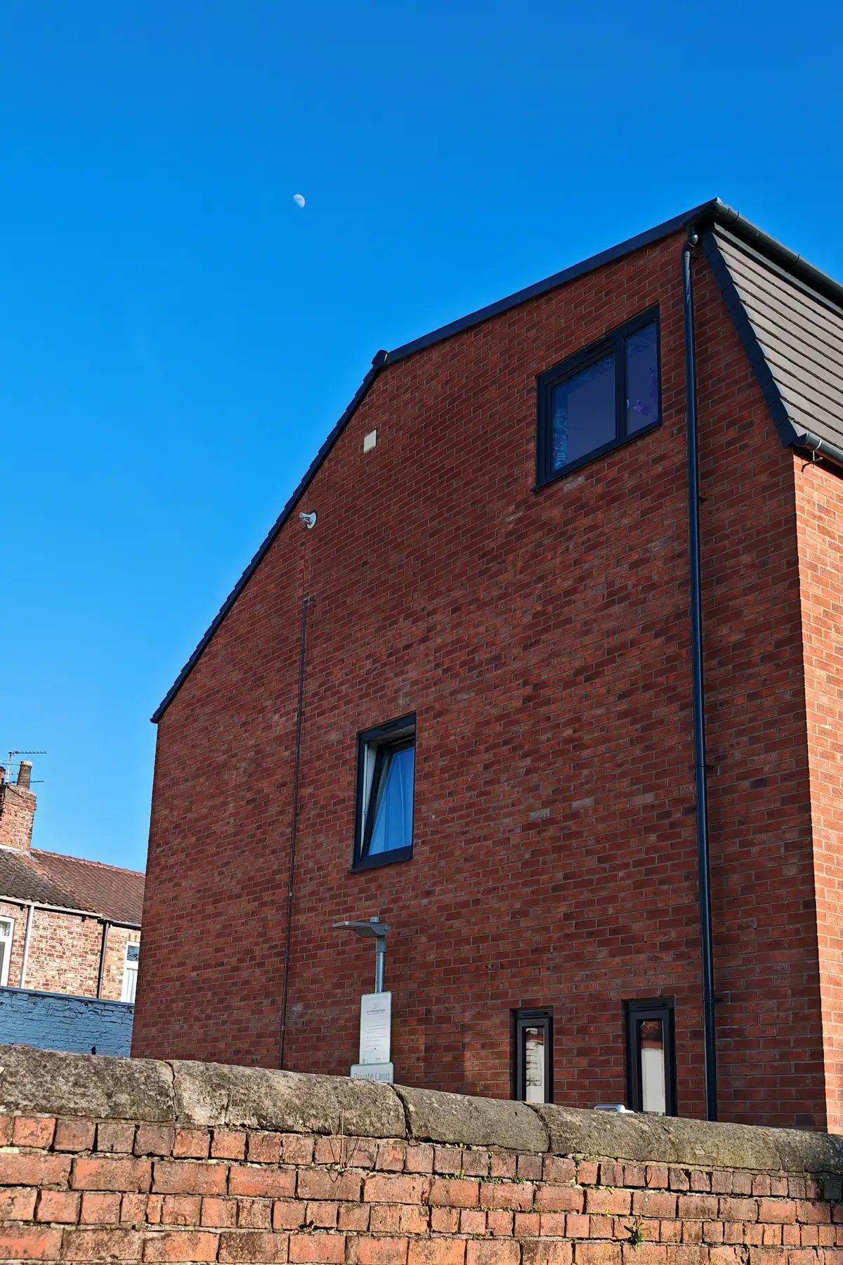 North gable elevation of Hawthorn Street Apartments viewed from the Hawthorn Street parking court, Layerthorpe York, with EV charging bollard in the foreground and the plain red-brick gable rising above the existing stone boundary wall on this brownfield infill site.