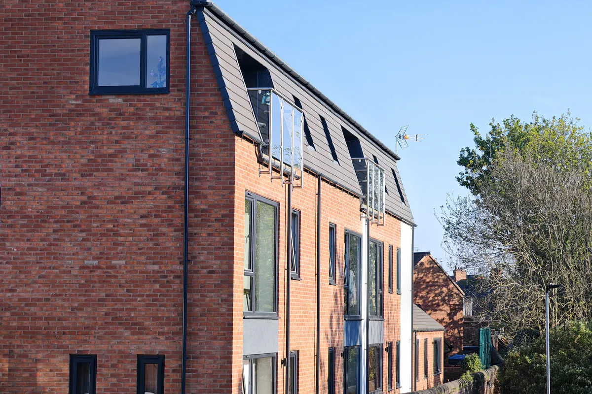 Corner view of Hawthorn Street Apartments from the Foss Islands cycleway, Layerthorpe York, showing the plain red-brick north gable end and west elevation with dark slate mansard roof and recessed dormer windows — contextual new-build architecture by Fining Associates.