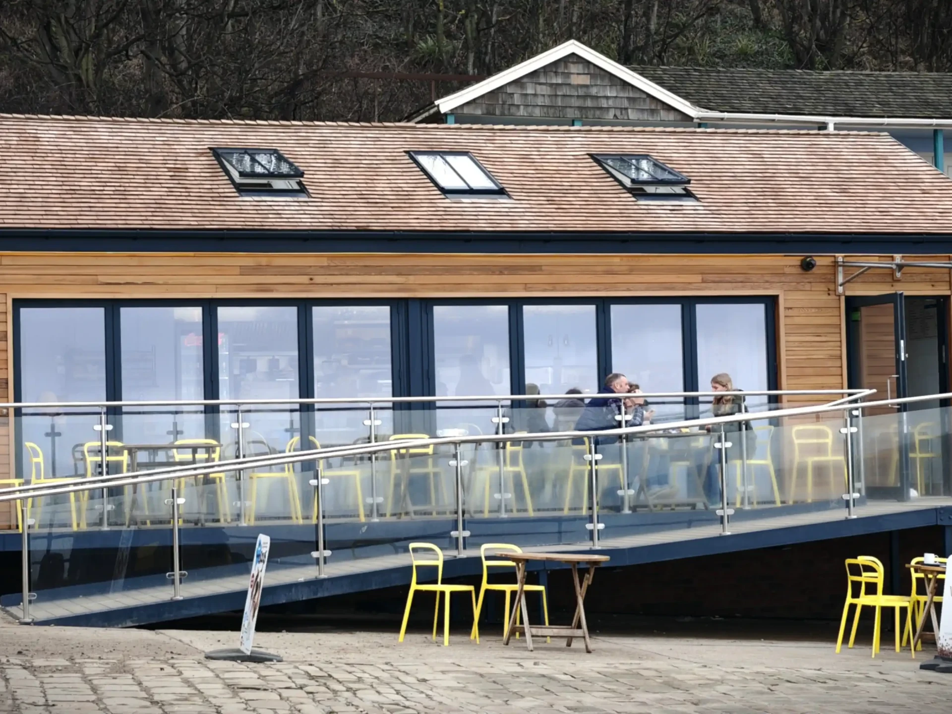 Full front elevation of the completed Baker's Snacks café at Coble Landing, Filey, showing the Western Red Cedar shingle roof, three conservation rooflights, horizontal timber cladding, large glazed bi-folding frontage, stainless steel and glass disabled access ramp and raised terrace, with the traditional wooden beach chalets visible to the right — Fining Associates Chartered Architects, York