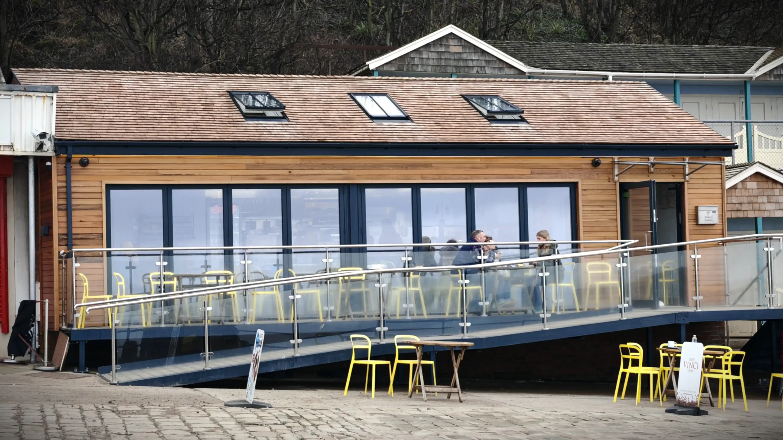 Full front elevation of the completed Baker's Snacks caf&eacute; at Coble Landing, Filey, showing the Western Red Cedar shingle roof, three conservation rooflights, horizontal timber cladding, large glazed bi-folding frontage, stainless steel and glass disabled access ramp and raised terrace, with the traditional wooden beach chalets visible to the right &mdash; Fining Associates Chartered Architects, York
