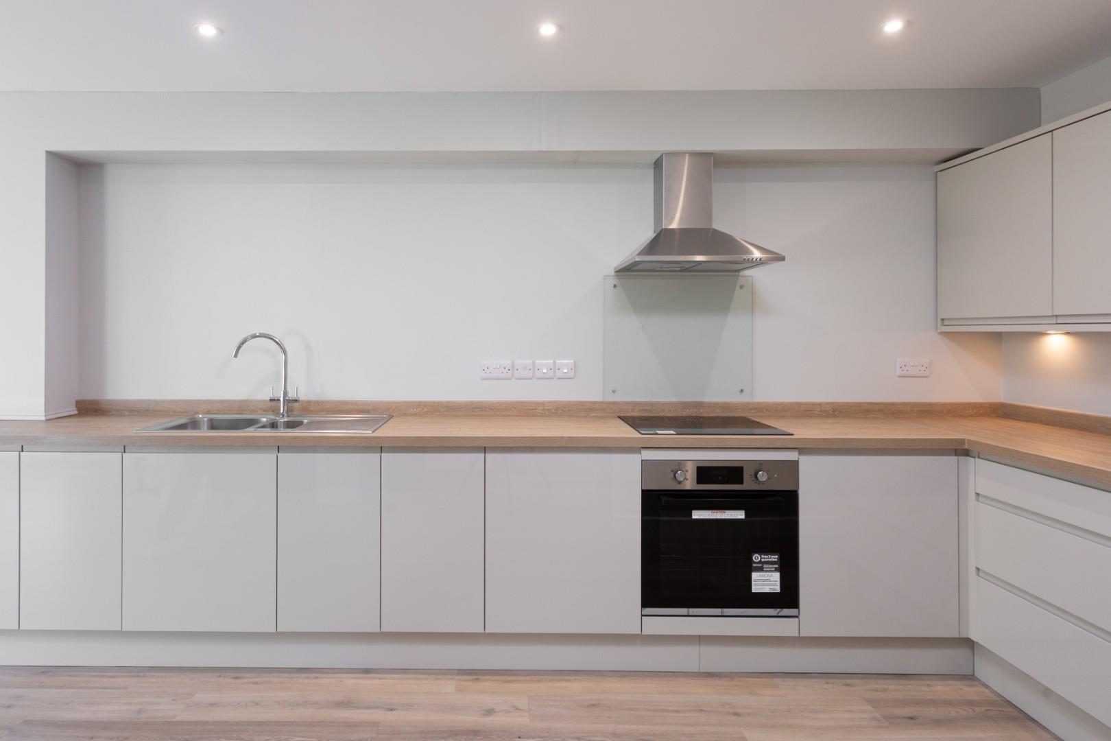 A bright, contemporary handleless kitchen interior forming part of the ground-floor living space in the new build backland and infill dwelling at Burton Stone Lane, York, designed by Fining Associates – Chartered Architects. Flat-fronted base units in a warm soft grey run in an L-shaped configuration beneath a continuous oak-effect laminate worktop. On the left, a stainless steel single-bowl inset sink with drainer and a chrome arc mixer tap sits beneath a generous expanse of plain white-painted wall. A black ceramic induction hob is flush-set into the worktop to the right, with a stainless steel pyramid extractor hood above and a clear glass splashback panel behind. A stainless steel and black glass built-under single oven sits below the hob. Matching handleless wall units with recessed under-cabinet LED lighting occupy the upper right corner. White double socket outlets run along the wall at worktop height. Recessed ceiling downlights illuminate the space from above, and pale oak-toned engineered wood plank flooring extends across the entire room.