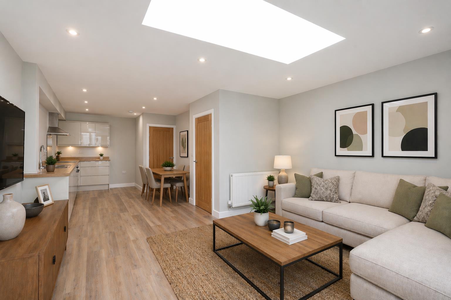 The ground-floor open-plan living, dining, and kitchen space of the new build backland and infill dwelling at Burton Stone Lane, York, designed by Fining Associates – Chartered Architects, photographed from the living room end looking towards the kitchen. Dominating the white ceiling, a very large rectangular flush-mounted rooflight floods the room with natural daylight from above — the primary daylighting device for this single-storey rear projection on a constrained urban site. In the right foreground, a cream L-shaped corner sofa dressed with sage-green and geometric-patterned cushions faces a rectangular oak coffee table on a jute rug, styled with stacked books, dark ceramic candle holders, and a potted fern; a cream-shaded table lamp and a small succulent sit on a round dark timber side table behind. Two abstract artworks in black frames with circular designs in sage, cream, and dark tones hang on the right-hand wall. A dark oak media unit and a large flat-screen television are partially visible at the left edge. At the centre of the room a dining table and taupe upholstered chairs define the dining zone, with two warm honey-oak panelled internal doors and a small framed artwork visible on the back wall. The L-shaped kitchen with gloss off-white units, under-cabinet LED lighting, and a stainless steel extractor hood is visible in the background. Pale oak-toned engineered wood plank flooring and soft sage-grey walls run throughout.