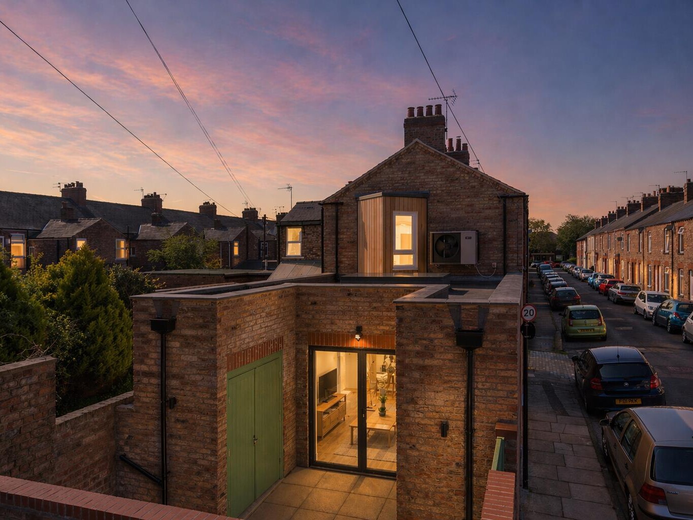 An elevated dusk exterior photograph of the completed new build backland and infill dwelling designed by Fining Associates – Chartered Architects, fronting onto Surtees Street at the rear of 175 Burton Stone Lane, York. The compact two-storey red brick building is photographed at blue hour, with warm amber interior light glowing through large black-framed glazed double doors at ground-floor level, revealing a furnished living room with television and timber furniture within. A green-painted door sits to the left of the entrance. Above the flat roof of the single-storey rear projection, a distinctive vertical timber-clad feature bay with a glazed window is illuminated at first-floor level, with an air source heat pump mounted to its right. The chimney stacks and gable of the parent building at No. 175 are visible rising behind. On either side, the tightly packed Victorian terraced rooflines of Surtees Street recede into the distance under a dramatic sky of pink, orange, and deep purple fading to blue — a 20mph sign and parked cars grounding the scene firmly in its urban residential context.