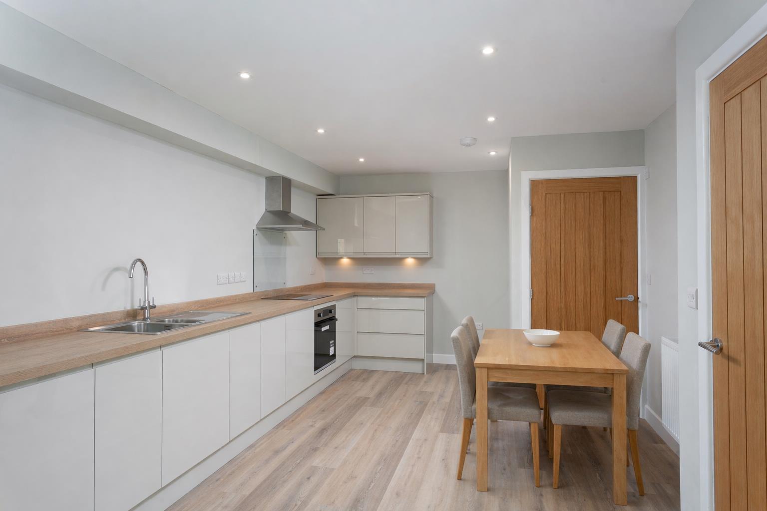 A light-filled open-plan kitchen and dining room interior in the new build backland and infill dwelling at Burton Stone Lane, York, designed by Fining Associates – Chartered Architects. The L-shaped kitchen runs along the left wall, fitted with flat-fronted handleless base and wall units in a warm off-white finish beneath a continuous oak-effect laminate worktop. A stainless steel single-bowl sink with drainer and a chrome arc mixer tap occupies the far left, with a black ceramic induction hob set into the worktop to the right, a stainless steel pyramid extractor hood above it, a glass splashback behind, and a built-under stainless steel oven below. Wall units with under-cabinet LED strip lighting wrap the far corner. To the right, a simple four-seater rectangular oak dining table with grey upholstered chairs and a single white ceramic bowl occupies the dining portion of the space. Two warm honey-toned panelled oak internal doors with chrome lever handles are set into the right-hand wall. Pale oak-toned engineered wood plank flooring runs throughout, and recessed LED downlights are distributed evenly across the white ceiling.