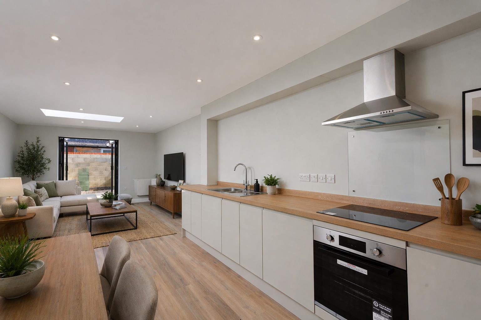 A wide-angle view of the full depth of the ground-floor open-plan kitchen and living space in the new build backland and infill dwelling at Burton Stone Lane, York, designed by Fining Associates – Chartered Architects, photographed from the kitchen end looking through to the courtyard beyond. In the right foreground, flat-fronted handleless base units in warm off-white support a continuous oak-effect laminate worktop with an inset stainless steel sink and chrome arc tap, a black ceramic induction hob, and a large stainless steel pyramid extractor hood above with glass splashback; a stainless steel built-under oven sits below. The space opens fully into a furnished living area where a cream corner sectional sofa with sage-green and neutral cushions faces a dark walnut media unit and flat-screen television. A jute rug, rectangular black metal-framed coffee table, ceramic table lamp, and a potted olive tree anchor the living zone. A dining table and upholstered grey chair are partially visible in the left foreground. A large rectangular flush rooflight in the white ceiling draws natural daylight into the centre of the single-storey plan from above. At the far end, black-framed glazed bifold doors open directly onto the private courtyard, with the grey concrete blockwork boundary wall and green-painted gate visible beyond. Pale oak-toned engineered wood plank flooring runs continuously throughout.